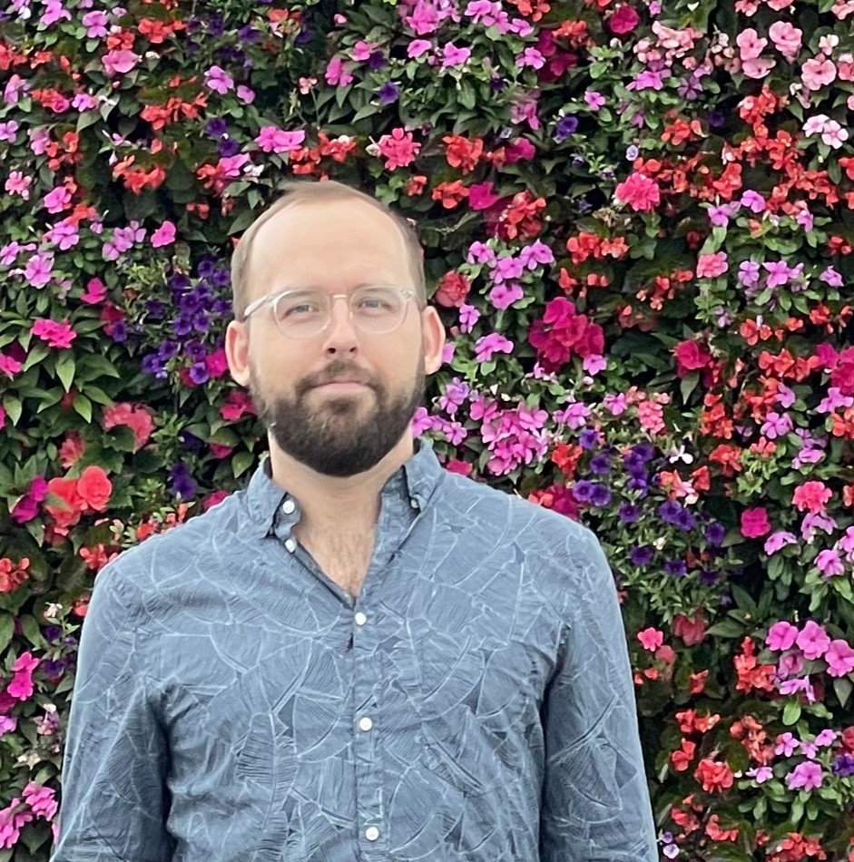 a picture of the candidate (white, mid-thirties, wearing clear glasses with a beard) wearing a blue botanical patterned shirt standing in front of a wall of flowers. 